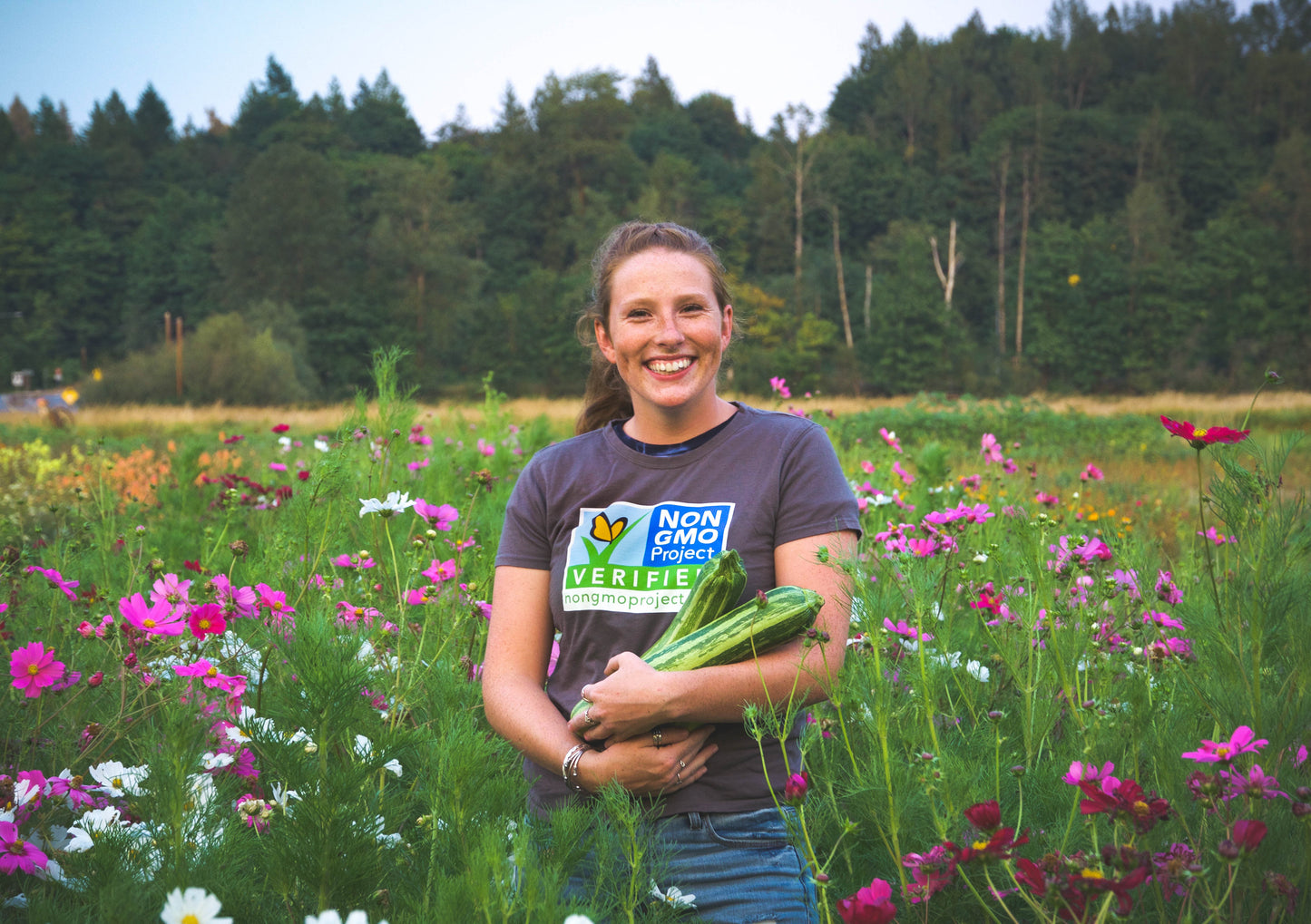 Woman standing in meadow, holding zucchinis and wearing Non-GMO Project gray t-shirt with large logo on chest