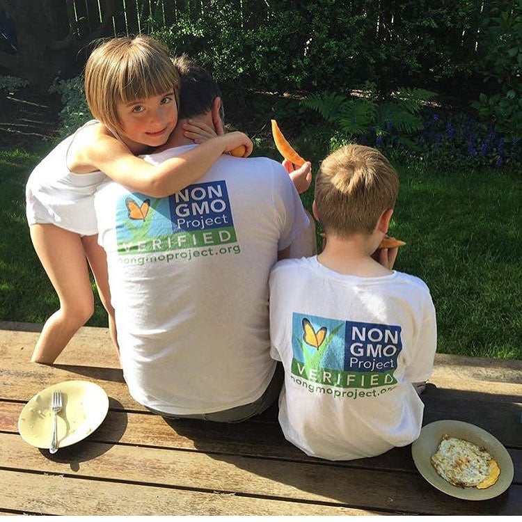 Man and children facing away , wearing Non-GMO Project t-shirts.  The back of the t-shirts show the Non-GMO Project logo and nongmoproject.org underneath