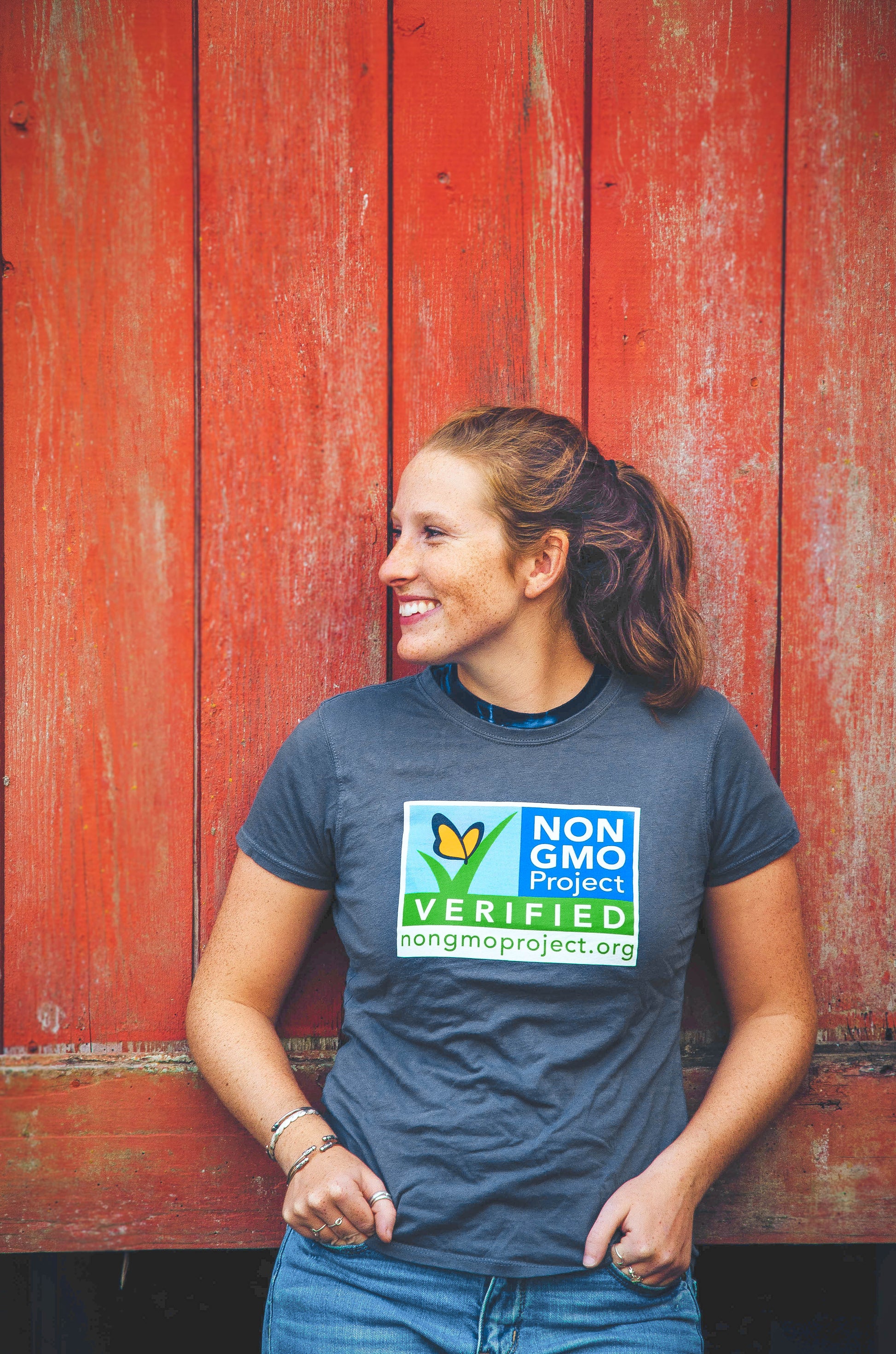 Woman standing in front of red barn wall, wearing Non-GMO Project gray t-shirt with front displaying  large logo on chest area