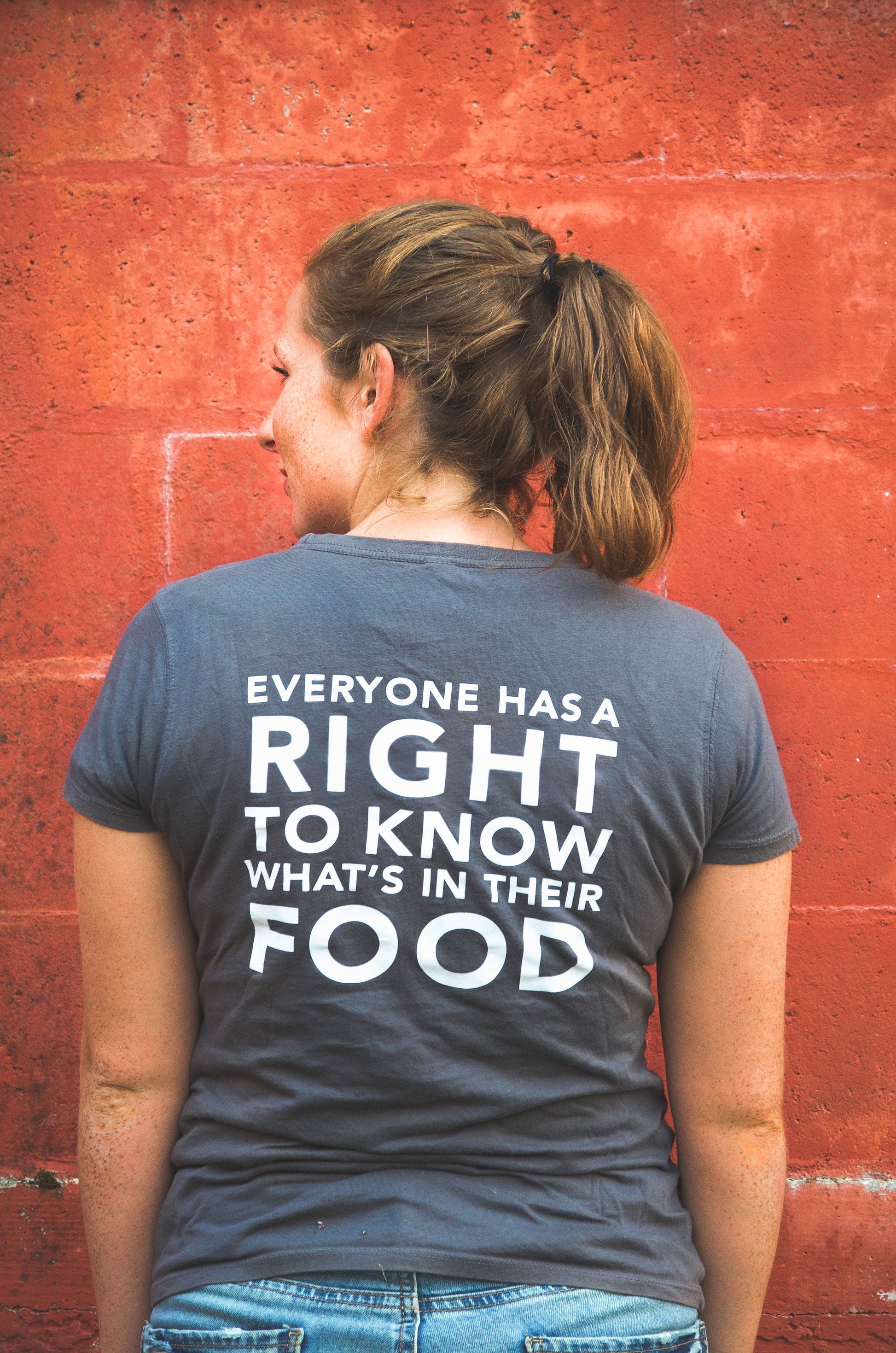 Woman standing in front of red barn wall, facing away from camera, wearing Non-GMO Project gray t-shirt with the back showing the quote "Everyone has a right to know what's in their food"