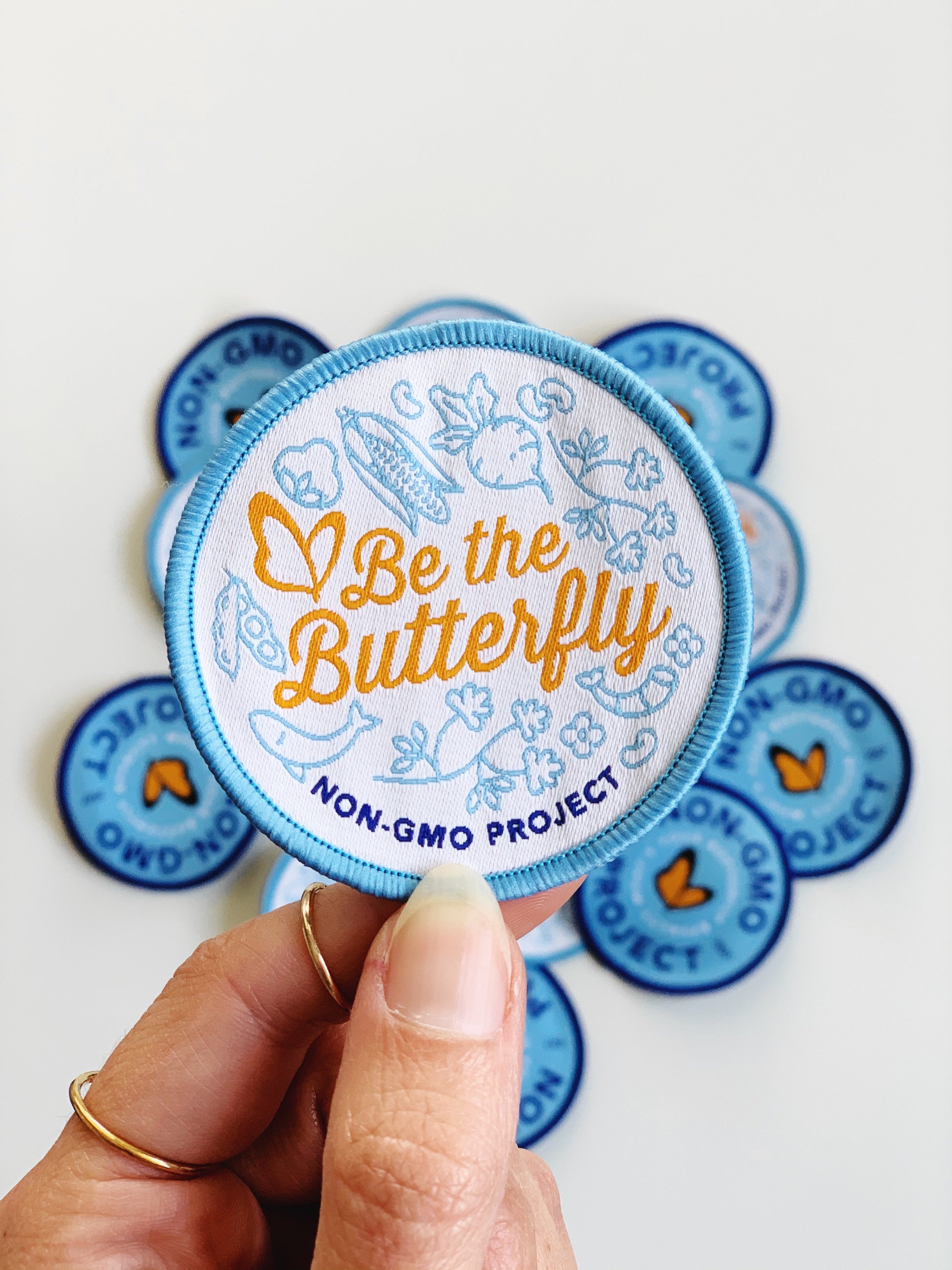 Non-GMO Project "Be the Butterfly" light blue, white, and orange woven patch, with a pile of patches in the background